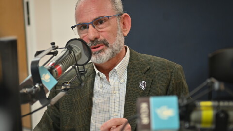 A man sits inside a radio studio. He is talking at a microphone and gesturing with his left hand.