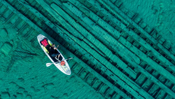 a woman on a paddle board looks over the side at a shipwreck below the green-blue water