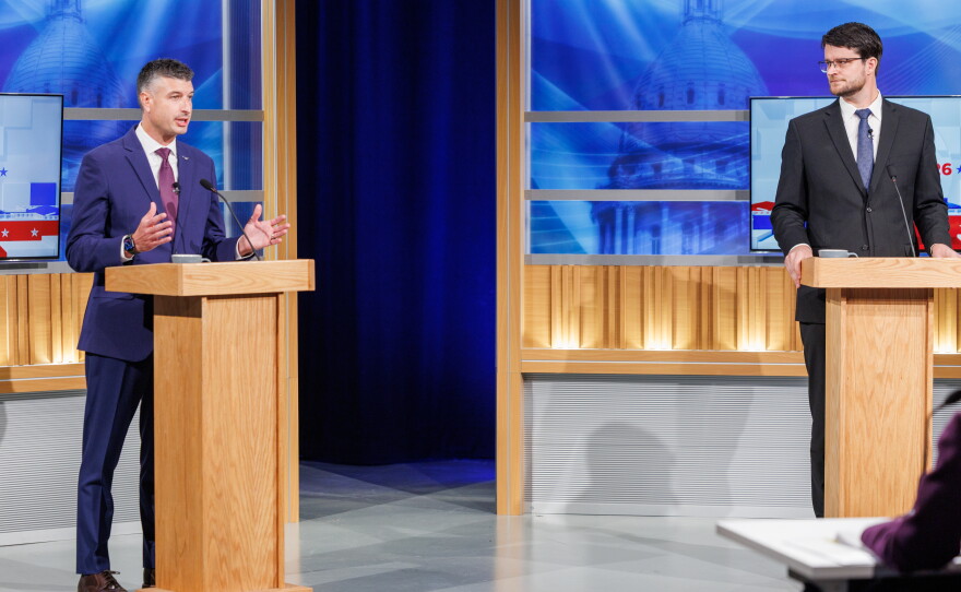 Two men in suits stand at podiums in a TV studio