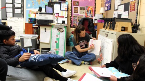 Three elementary schoolers sit on bean bag chairs. They are gathered around a teacher who is drawing on a white board.