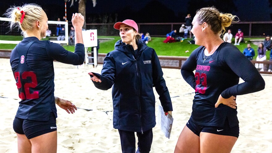 a beach volleyball coach in a black nike jacket and long pants carrying a clipboard talks to two players in black tops with red numbers and b lack shorts on a sandy beach volleyball court