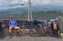 Smiling woman in shorts sitting on bench with graffiti. Two biked leaning against the bench filled with rucksacks. Mountains in background