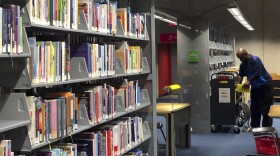 Books fill shelves at a library. In the background, a man reshelves books.