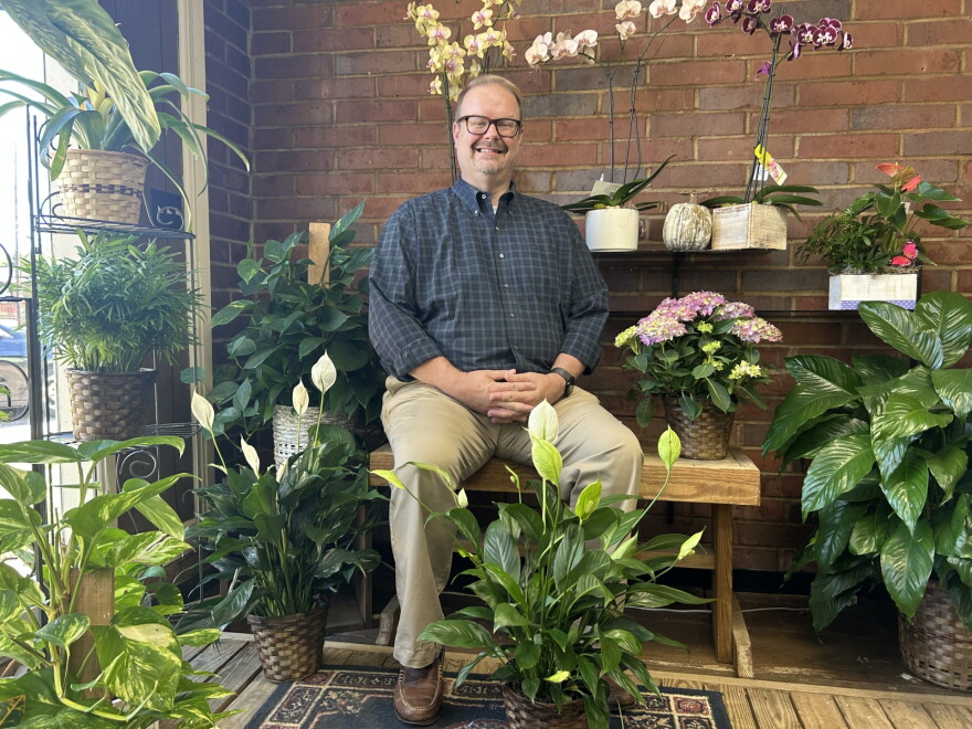 A smiling man with glasses, wearing a button-down plaid shirt and sitting on a bench surrounded by plants.