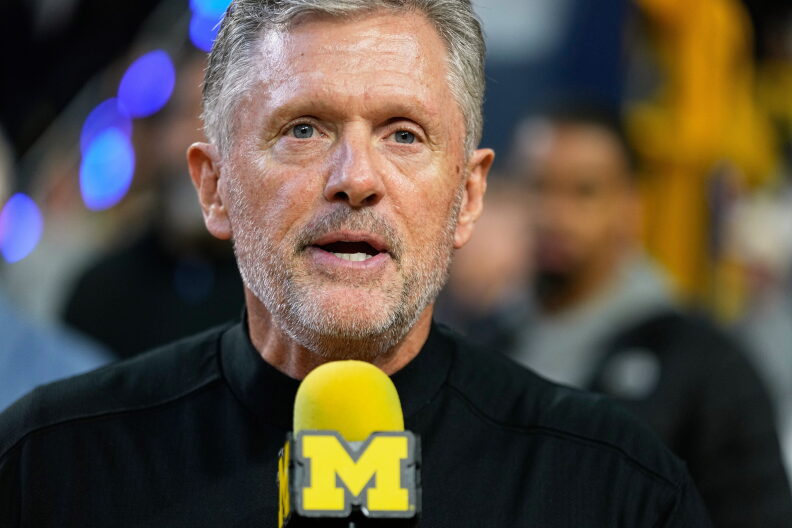 A man with gray hair and a beard speaks into a yellow microphone with a blue “M” logo, with people and blurred lights in the background.