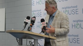 Sorensen wears a tan suit and blue shirt and stands at a podium. 