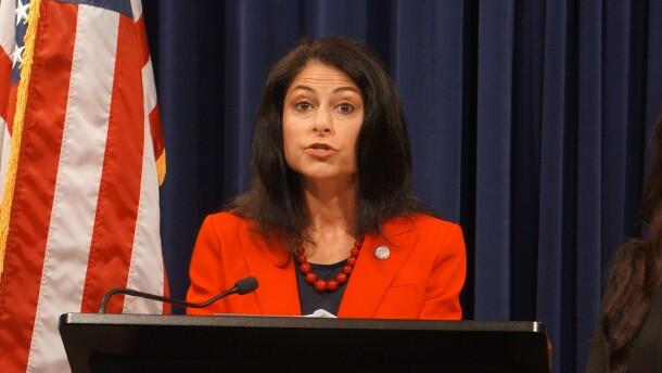 Dana Nessel, a white woman with dark brown hair and wearing a read blazer, stands at a podium in front of a dark blue backdrop and United States flag.