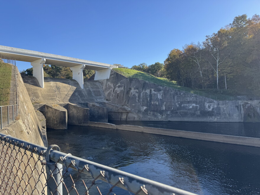 A concrete dam with a bridge atop of it spans a river, with trees on the far bank.