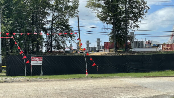 A black fence with a sign that reads "Private Property -- No Trespassing" stands in front of a property that contains smoke stacks, shipping containers and other industrial equipment. 