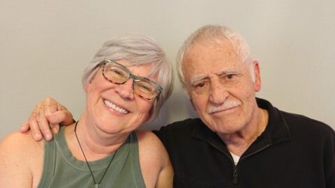 Kay and Arnold Martinez share a sideways hug at their StoryCorps appointment in St. George, Utah.  Kay has short white-gray hair and wears glasses with colorful frames, blue and black on the top tan and black on the bottom.  She smiles with one arm under her father's arm. She is wearing an army green tank top, and a large green pendant hanging from a dark cord around her neck.  Arnold has one hand on Kay's shoulder, and his thumb is holding his index finger.  Arnold has white hair and mustache and wears a black  pullover with a short zipped placket, a left chest pocket with a button, an da piece of paper  slightly extending from the pocket.  Arnold looks toward the camera in a matter-of fact way, his dark eyes expressing love, and his mouth in a straight line.