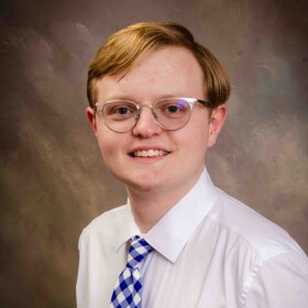 Nicholas Hazelett. A young, red-haired man wearing glasses and dressed in a white dress shirt and blue checkered tie.