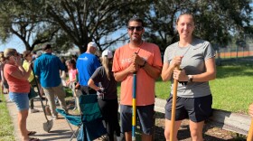 Man and woman stand with shovels in a line of people at a sandbag filling station.