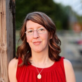 Bobi Conn wearing red top and glasses, leaning against wood post.