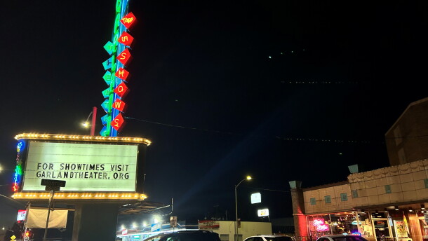 Cars sit parked near the front entrance of the Garland Theater, with the theater's sign lit up in colorful lights. 