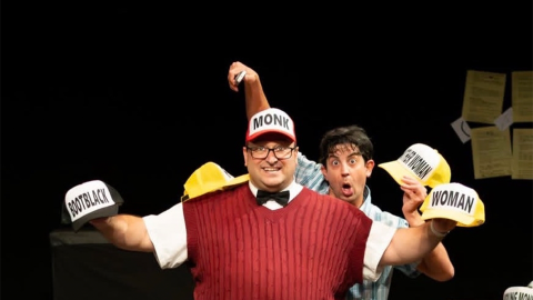 A man in a red sweater vest, a black bowtie, and khakis wears a red trucker hat that says "Monk" and holds two other trucker hats. Behind him, a man in a short sleeved plaid button up holds another trucker hat in one hand and a remote in the other. 