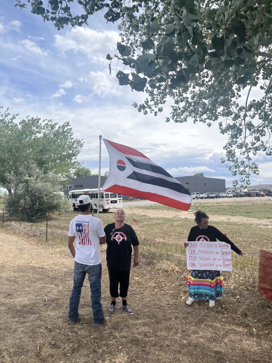 A boy in an American flag t-shirt holds up an Arapaho flag. He stands next to an older woman in a black t-shirt and another woman in a colorful skirt holding up a protest sign.