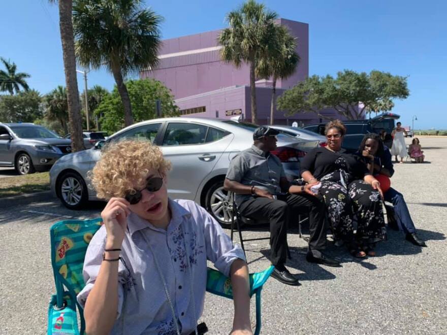 Boy with curly blond hair and sunglasses sits in a lawn chair and looks at camera