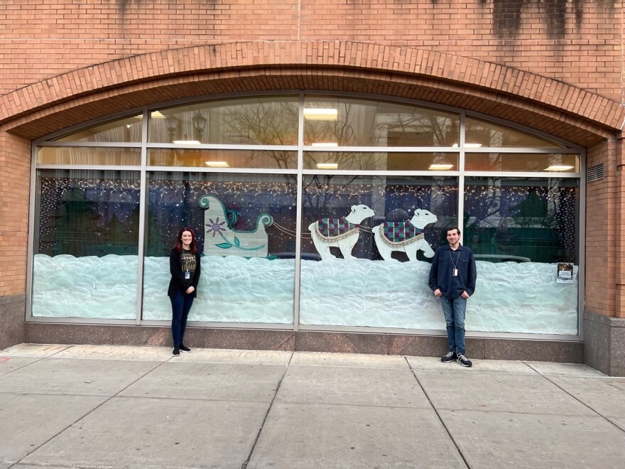 Two people stand on each side of a wide window display of two polar bears pulling a sleigh through snow.