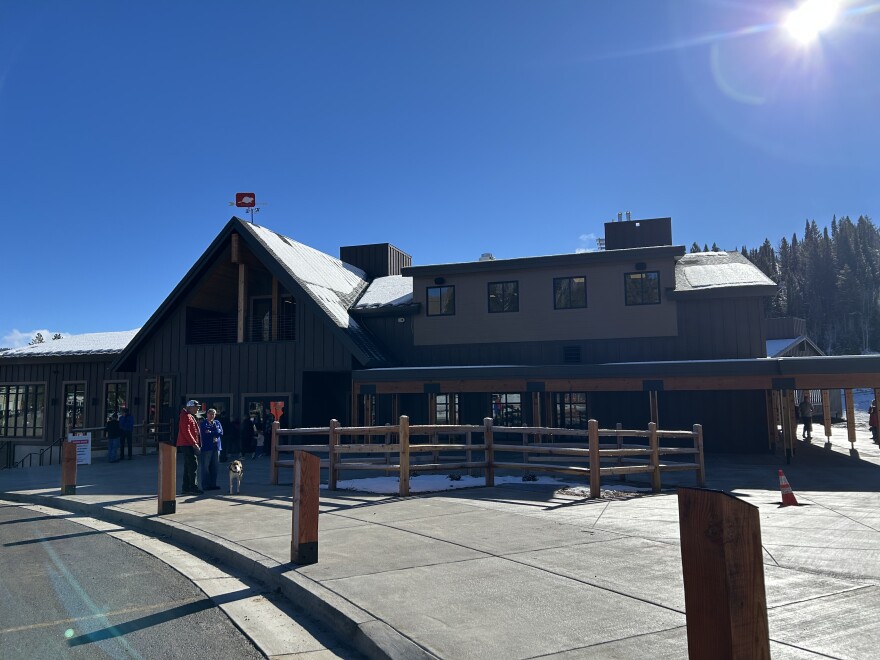 A large brown building stands in front of some pine trees