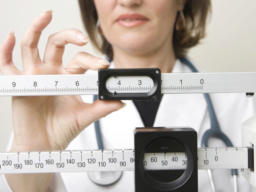 Doctors office scale in the forefront with a female doctor standing in the background. The doctor is in a white coat with a stethoscope draped around her shoulders  with her fingers on the scale weight adjustment slides. 