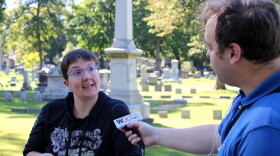 A man with a microphone interviews a woman in a cemetery. 