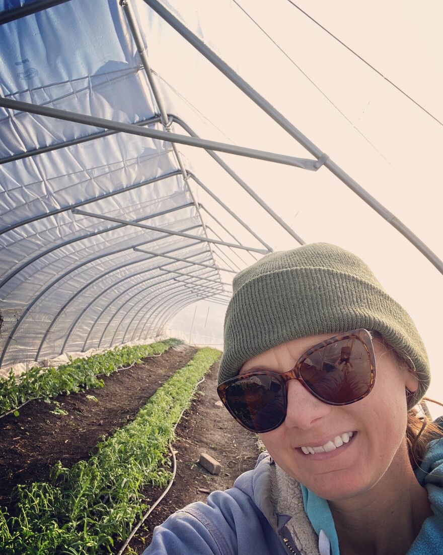 A woman in a beanie smiles in front of crops planted inside a farm structure.