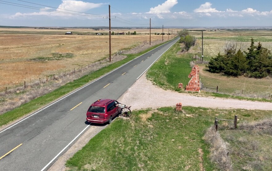 A red minivan is seen parked along a stretch of a rural, paved road in this aerial image. A hand is reaching into a mailbox from the passenger side of the vehicle. The grassy fields are mostly yellow and brown, but some areas are greening with spring. Letter carrier Roger McDonald delivers mail to a ranch on May 9, 2025, in rural Gering, Nebraska. McDonald’s route is nearly 150 miles long and contains 334 delivery points. The long distances and low population density are often blamed in part for the annual losses reported by the Postal Service.