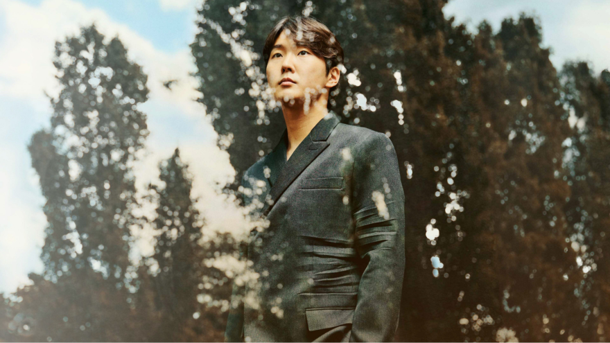 Seong-Jin Cho stands at a window with his hands in his pocket, overlooking a forest.