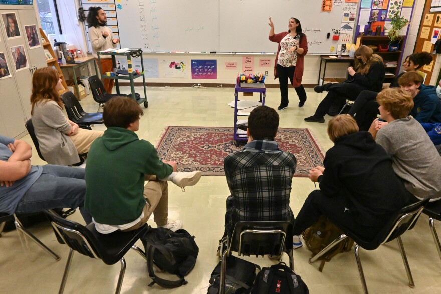 Several students sit in a semi-circle on chairs listening to a teacher who is talking in front of a large dry-erase board in a classroom.