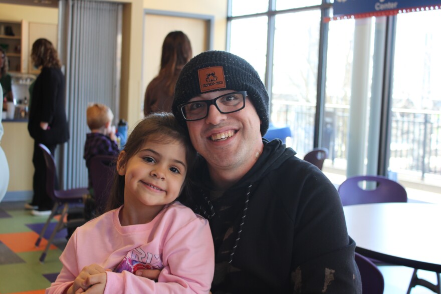 A man in a beanie and hoodie sits with a young girl on his lap in a pink sweater. Both are smiling. 