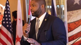 Black man with a beard and dark suit speaking into the microphone, looking to the left and an American flag to the left behind him