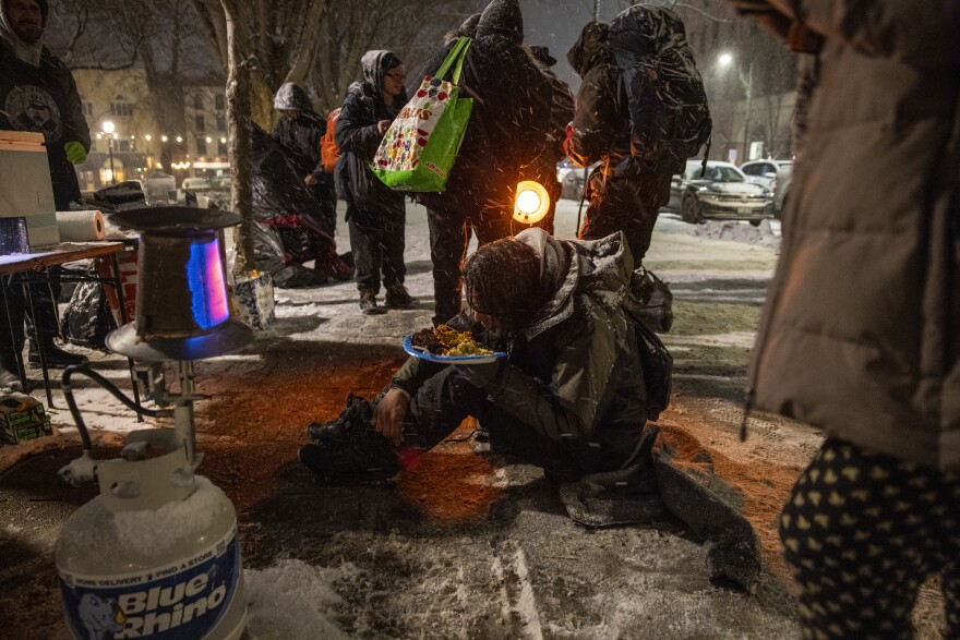 Snow piles up on a man with a plate of food. He sits on the sidewalk amid a handful of other people, who are standing. There are propane heaters set up nearby.