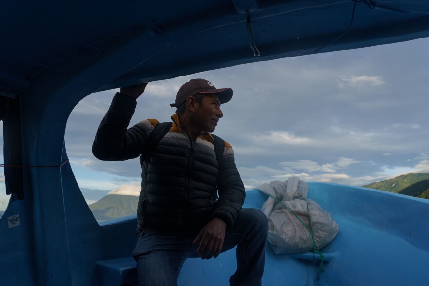 A man rides on the front of a public Lake Atitlán water taxi from Panajachel to San Pedro la Laguna at dawn. Behind him, the San Pedro volcano is visible. Lake Atitlán, Sololá, Guatemala.