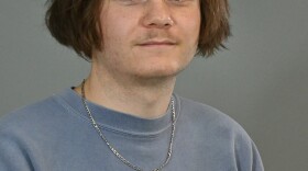 A young man wearing blue sweatshirt standing against a gray background. He is sort of smiling at the camera.