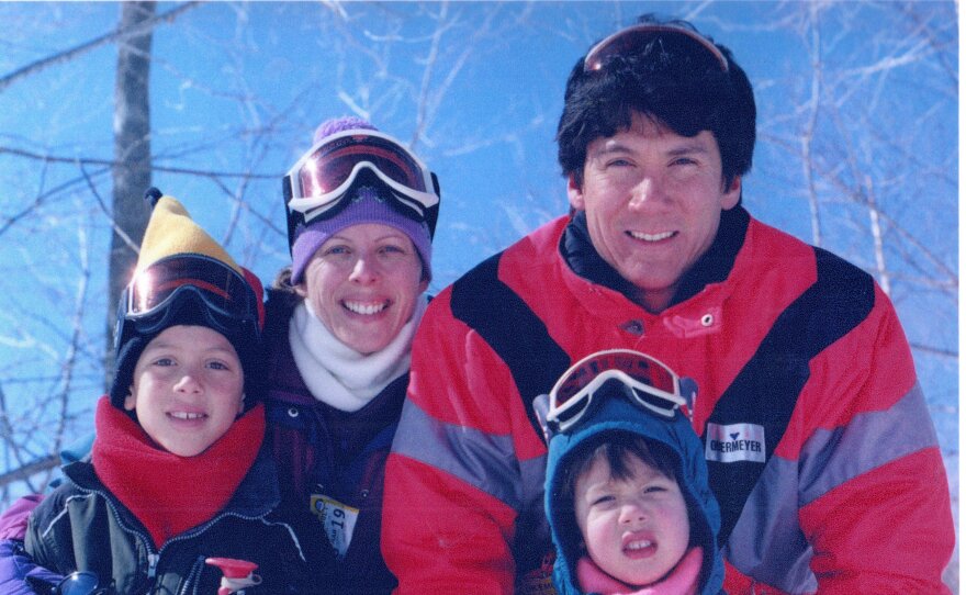 A photo of a young family, two children and two parents, wearing 1990s ski apparel, stand against a backdrop of bluebird sky and bare trees.