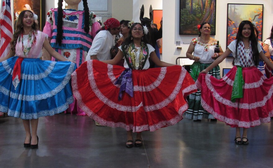 Three young women standing in a line hold up the sides of their large, brightly colored skirts and smile at the crowd