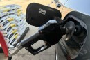 A black gas pump nozzle is inserted into a gray SUV with a pallet of bags of ice melting compound visible in the background. 