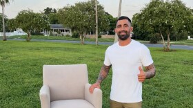 Man in white t-shirt stands outside next to a chair 