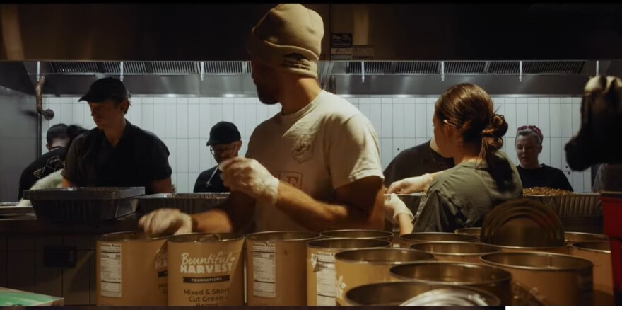 Several people stand in a food prep room, wearing hats and gloves. They are assembling free meals for a Thanksgiving event.