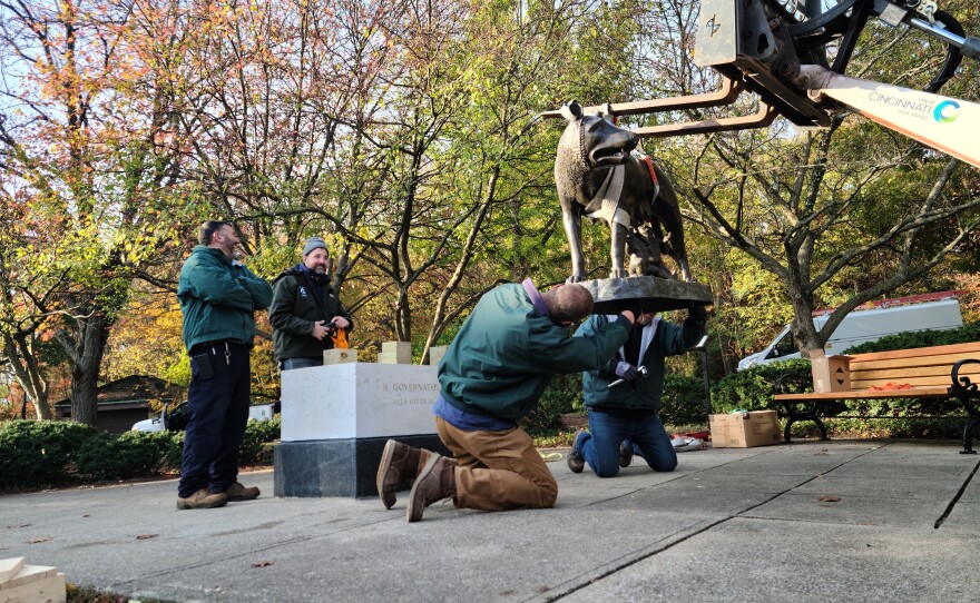 men screw bolts into the base of a statue held aloft by a forklift