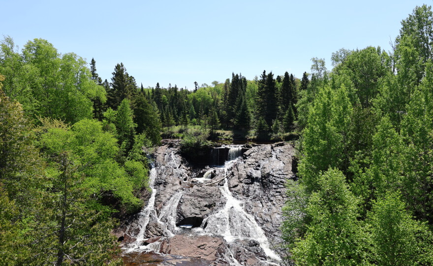 A picture of a waterfall flowing over dark basalt rock in Eagle River Falls, MI. It's surrounded on either side by green trees. 
