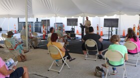 People sitting in chairs, facing Hudson, who is at a podium speaking with a screen behind him, all over a large tent the people are under.