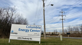 A white sign with blue lettering reads "Columbia Energy Center." The sign stands on green grass. A road and power lines are visible in the background. 