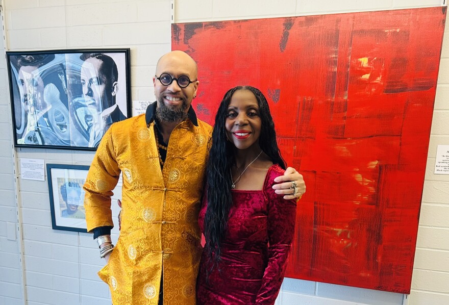 A man in a yellow jacket and woman in a velvet dress stand shoulder-to-shoulder in front of a big red canvas. 