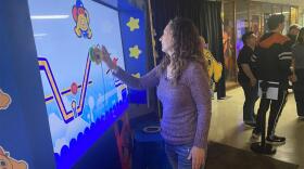 Carrie Principe playing the interactive Path Pursuit game on a large digital screen at Imagine RIT, with colorful cartoon graphics and other visitors watching nearby.
