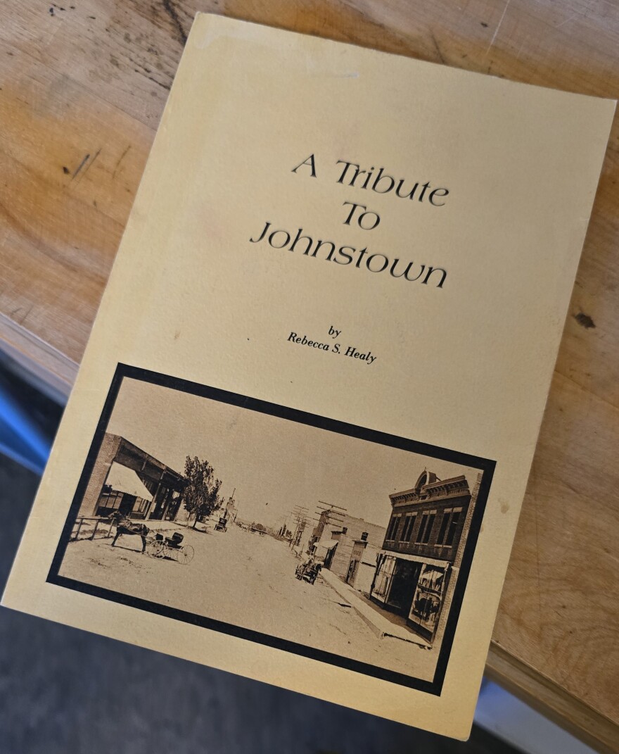A small, tan-colored book sits on a wooden table with a medium finish. The front cover of the book bears the words "A Tribute to Johnstown by Rebecca S. Healy" in black text. A black-and-white photo features Johnstown's early downtown area. Horse-drawn carriages travel on a dirt road lined with shops.