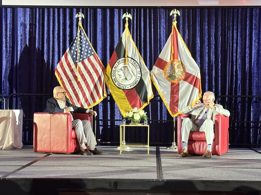 Two man in suits sit on stage in front of a display of flags