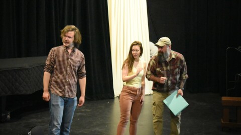 Three people standing on a black stage with a large white curtain behind them.