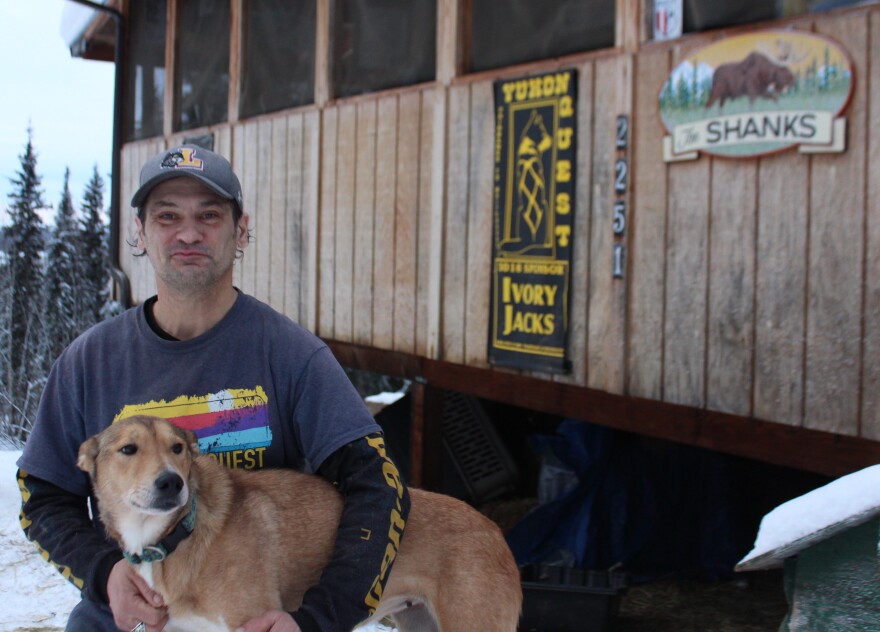 A man holsds a reddish dog in front of a log house