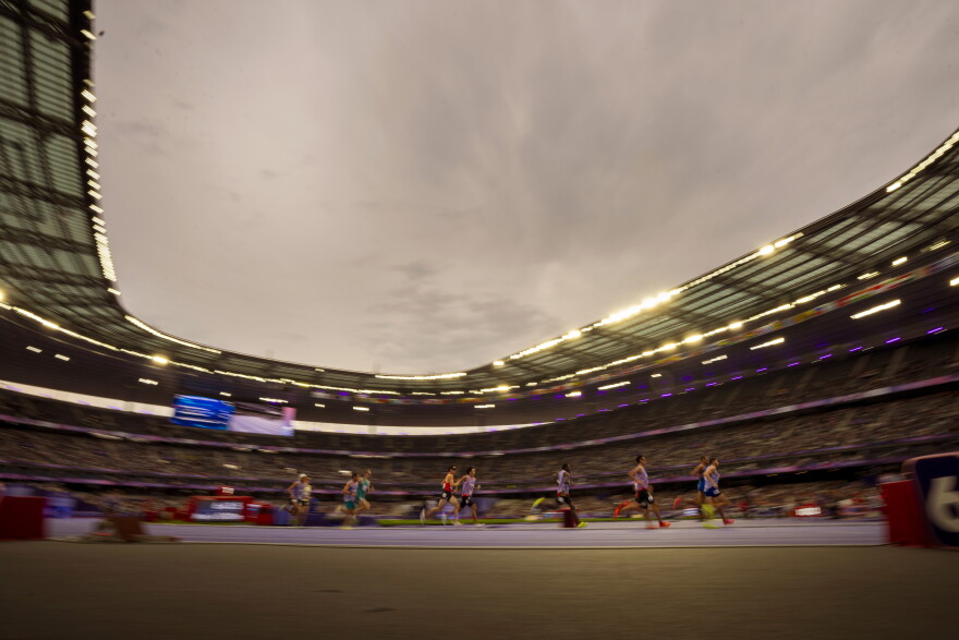 An artistic, slightly blurry image of athletes running in a lighted stadium. The athletes, from far away, are mostly in focus, while the background is slightly blurry. The stadium has an open room and gray skies can be seen above as the lights start to come on in the stadium. 